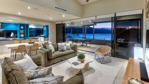 A modern living room with a large window overlooking a scenic lake, a dining area with a view, and a kitchen with blue accents. - Edge Apartment 7, Edge Apartment 7 by HIHA (Hamilton Island)