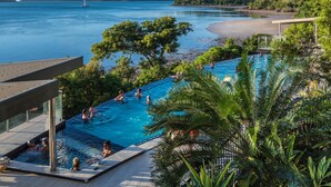 A pool area with people swimming, surrounded by lush greenery and overlooking a beach and water. - Edge Apartment 7, Edge Apartment 7 by HIHA (Hamilton Island)