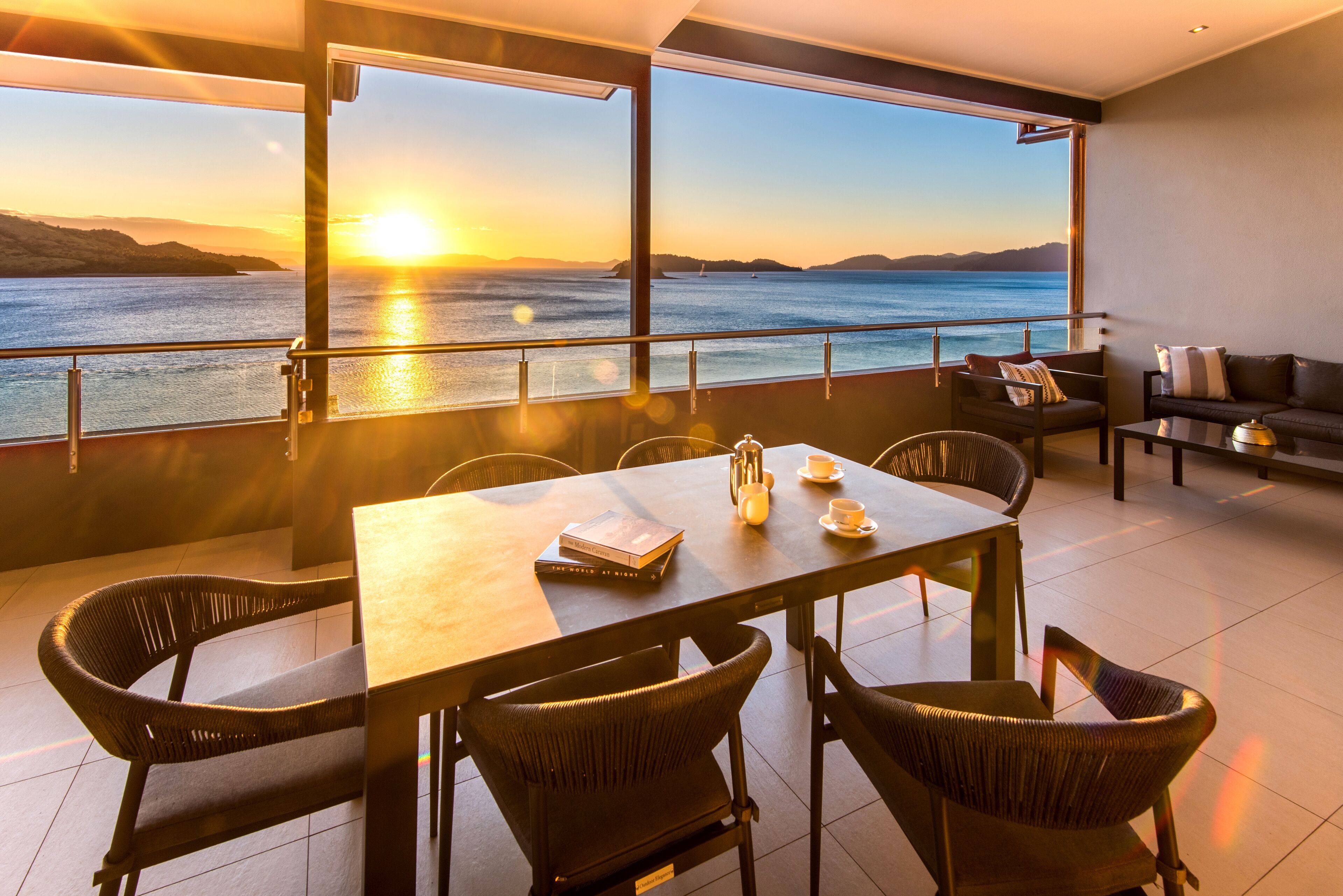 A modern dining area with a view of the sunset over the water, a table with chairs, and a sofa.