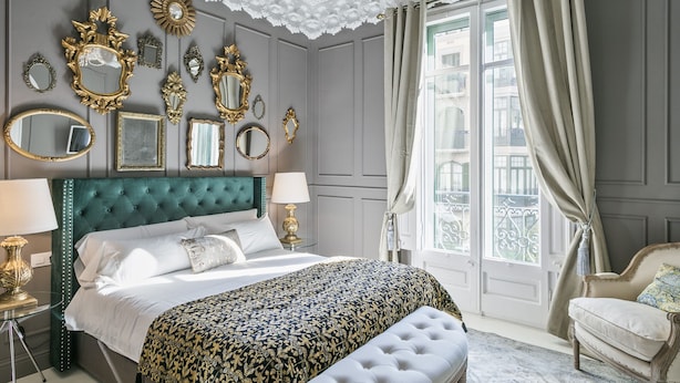 A bedroom with a large bed, multiple ornate mirrors, a tufted headboard, and a view of the city through large windows.