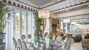 A spacious living room with a glass dining table surrounded by chairs, a chandelier, and potted plants.