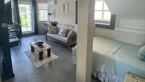 Un'ampia zona giorno con divano, tavolino e tavolo da pranzo. Presente una ventola a soffitto e una finestra che lascia filtrare luce naturale.