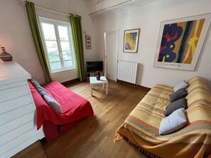 A room with a bed, a sofa, a small table, a radiator, and artwork on the walls.