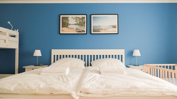 Een slaapkamer met een wit bed, twee nachtkastjes met lampen, een stapelbed links en twee ingekaderde foto's aan de muur.