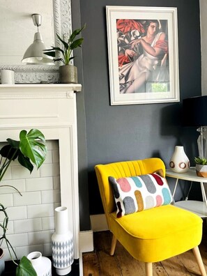 A room with a yellow armchair, a white fireplace, and a framed painting on the wall.