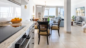 A modern kitchen with a marble countertop, a dining area with wicker chairs, and a view of the sea through large windows.