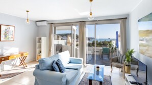 A modern living room with a sofa, a glass coffee table, and a TV. There is a balcony with a view of the sea.