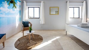 A room with a beach mural, two windows with curtains, a wicker chair, a small table with a plant, and a bed with towels.