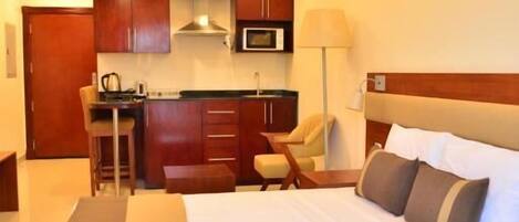 A hotel room with a bed, a desk, a chair, a microwave, and a kitchenette.