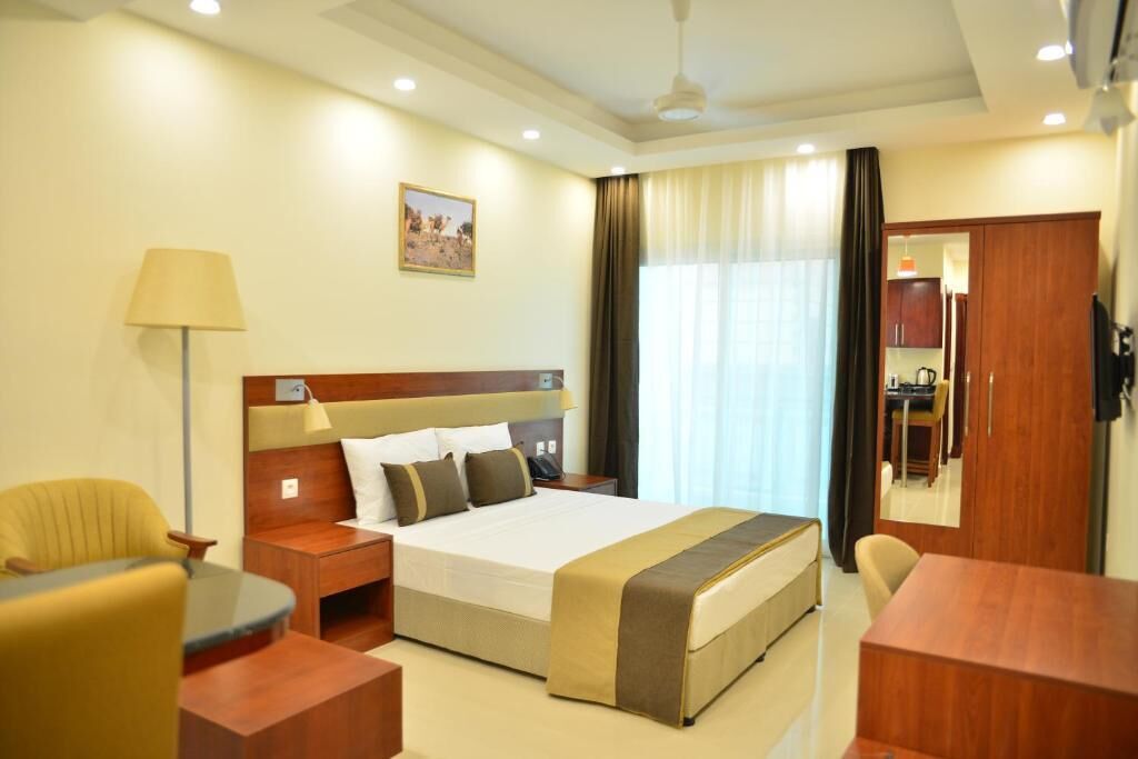 A hotel room with a large bed, a desk, a chair, a lamp, a painting on the wall, and a view of the dining area.
