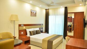 A hotel room with a large bed, a desk, a chair, a lamp, a painting on the wall, and a view of the dining area.