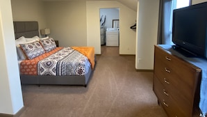 A bedroom with a bed, dresser, TV, and a laundry area in the background. - Chehalis upstairs 2 bd 2 bath apartment (Chehalis)