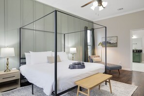 A four-poster bed with a wooden bench, a nightstand with a lamp, and a view of a kitchen through an open door.