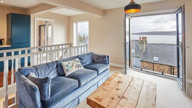 A modern living room with a blue sofa, wooden coffee table, and a balcony with a view of the sea. - Host & Stay | Watch House (Scarborough)