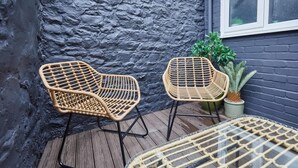 Two wicker chairs with metal frames on a wooden deck, a small potted plant, and a window with white frames.