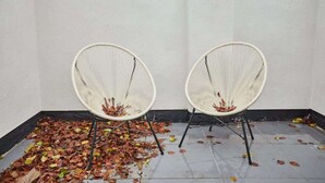 Two white wireframe chairs with black legs, placed in a corner with a white wall and a floor covered with fallen leaves. - Host & Stay | High Street Cottage (Norton)