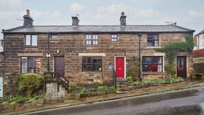 Une maison en pierre à deux étages, avec une porte rouge, un petit jardin à l’avant et un escalier apparent menant à l’entrée.