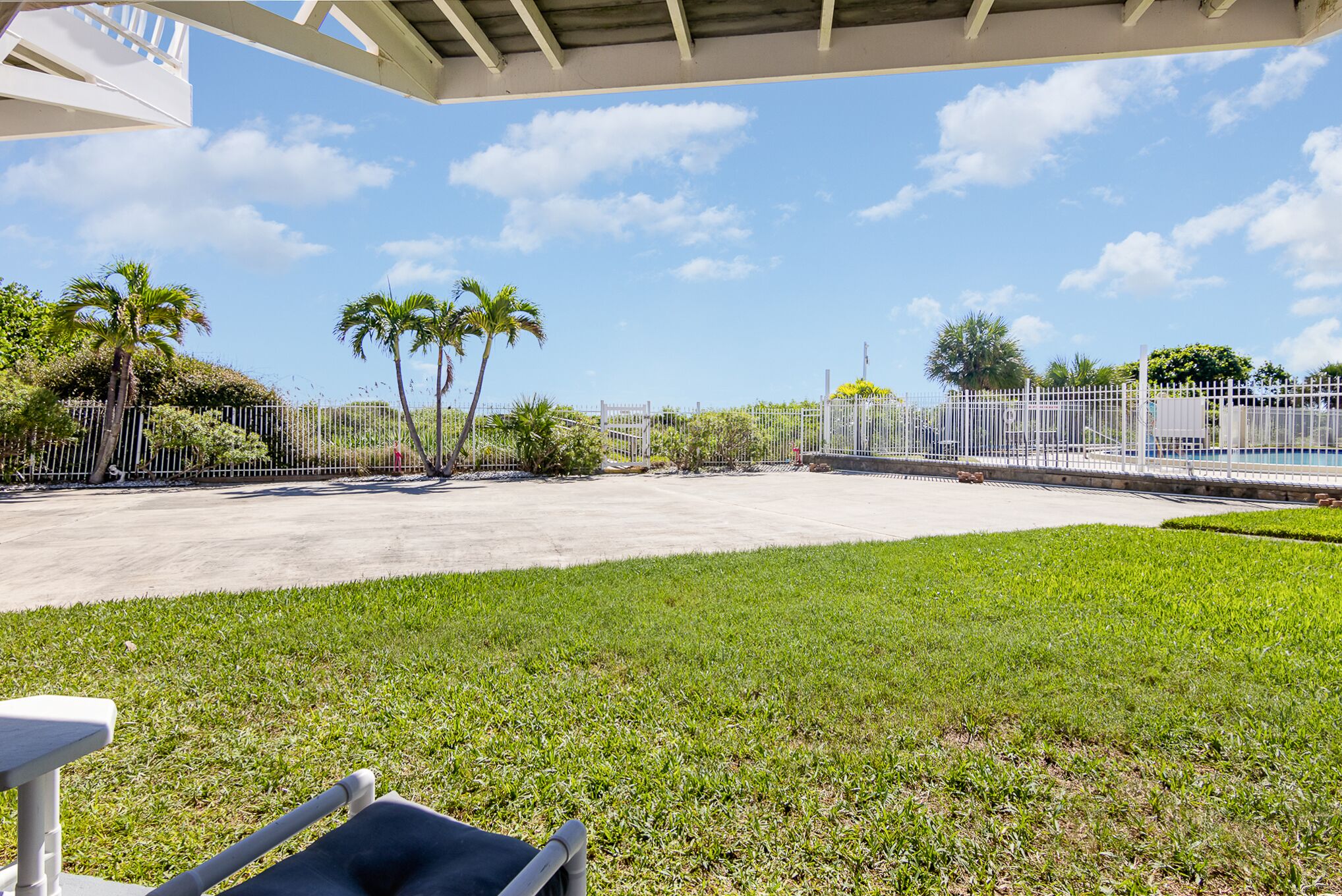 A covered outdoor area with a concrete floor, surrounded by a white fence, palm trees, and a grassy lawn.