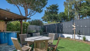 Un'area patio con tavolo e sedie in legno, circondata da un giardino recintato con erba sintetica e alberi.