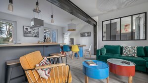 Un soggiorno moderno con un divano verde, una poltrona gialla e un poggiapiedi blu. Si intravede in secondo piano una zona cucina con una grande finestra.