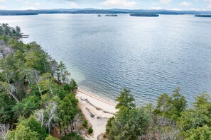 A coastal view with a sandy beach, dense forest, and calm water. - Wildwood on Lake Winnipesaukee Pet Friendly Beach Access Home Sleeping 10 (Moultonborough)