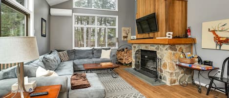 A modern living room with a stone fireplace, grey sofas, and a large window overlooking a forest.