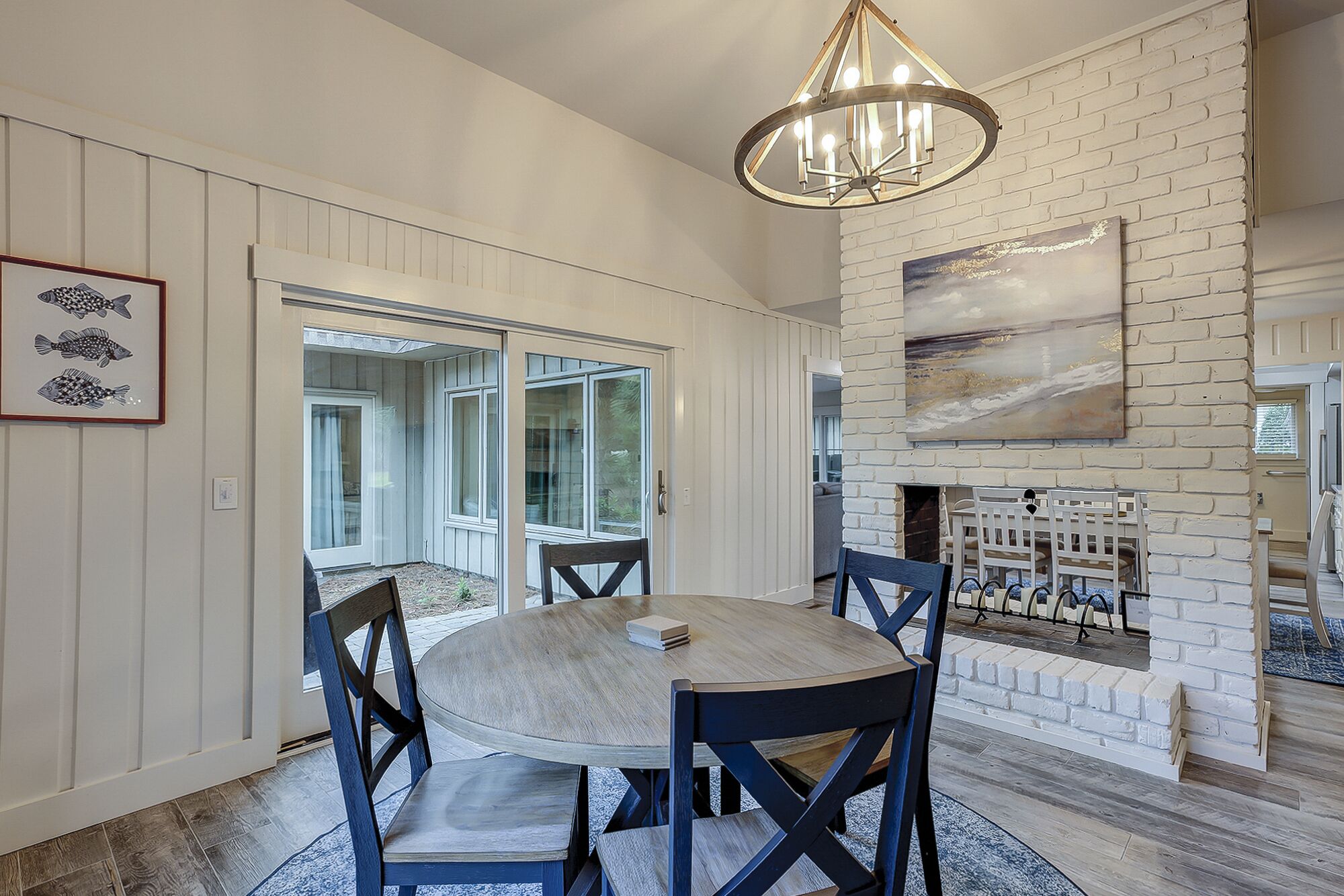 A dining area with a round table, chairs, and a chandelier. There is a fireplace with a brick wall and a painting above it.