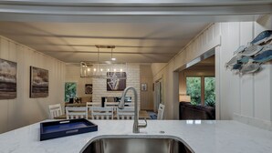 Una cocina moderna con encimera de mármol blanco, pileta de acero inoxidable y horno integrado. Hay obras de arte enmarcadas en las paredes y una zona de comedor con sillas blancas.