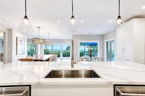 Une cuisine moderne dotée d’un grand îlot central, d’appareils électroménagers en acier inoxydable, de luminaires suspendus et offrant une vue sur l’extérieur.
