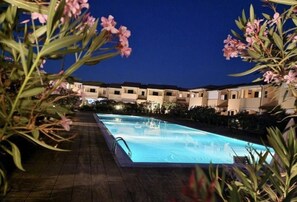 Une piscine dotée d’une terrasse carrelée, entourée d’immeubles avec des balcons, le tout sous un ciel étoilé.