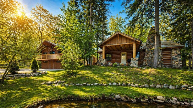 Une cabane avec une cheminée en pierre, une terrasse en bois et un étang devant.