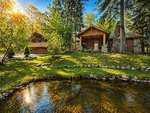 A cabin with a stone chimney, a wooden deck, and a pond in front.