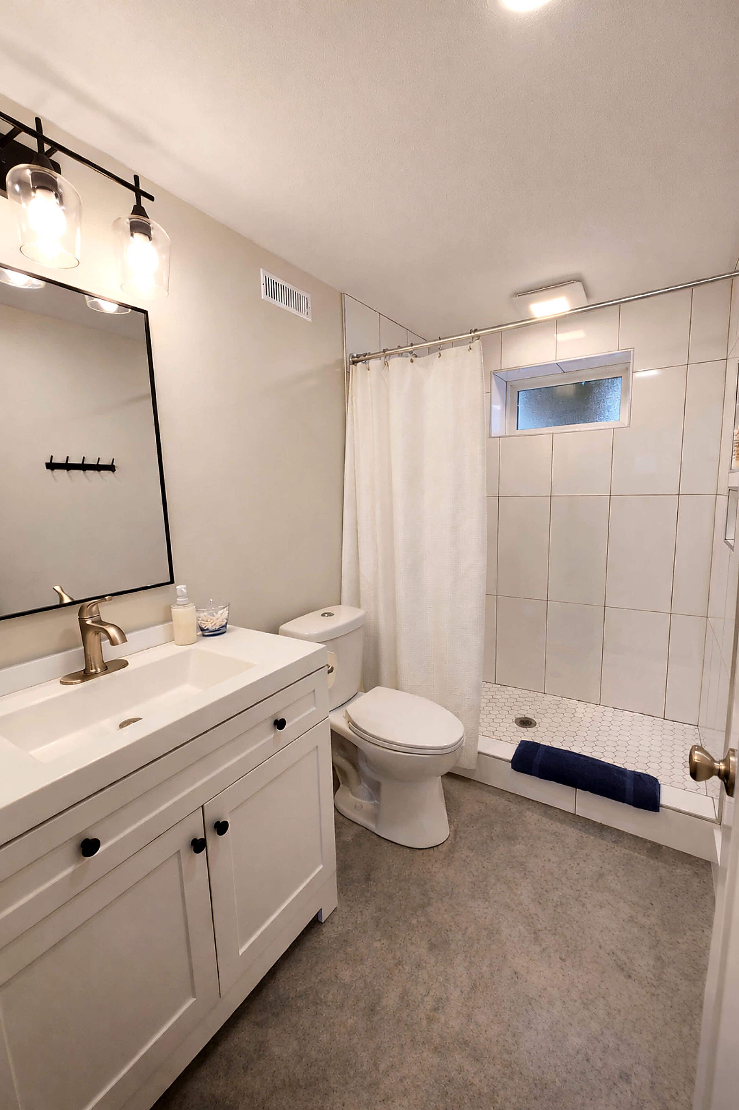 Un bany amb un lavabo blanc, un mirall, un vàter i una dutxa amb porta de vidre.