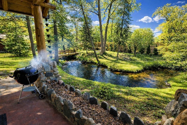 A barbecue grill set up near a pond with a wooden bridge, surrounded by lush greenery and trees.