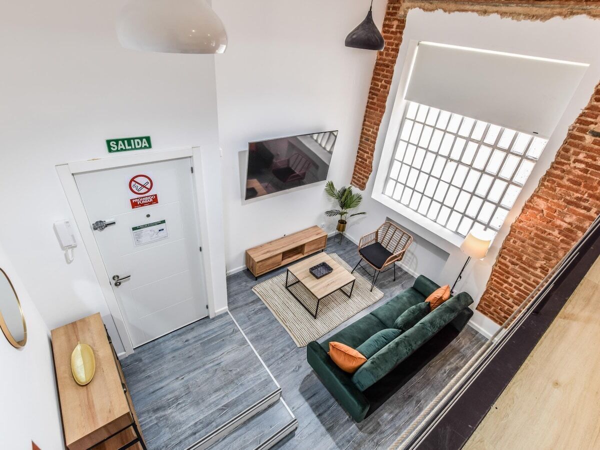 Un salon moderne avec un canapé vert, une table basse et un téléviseur fixé au mur. On y trouve une porte portant l’inscription « SORTIE » et un panneau indiquant qu’il est interdit de fumer.