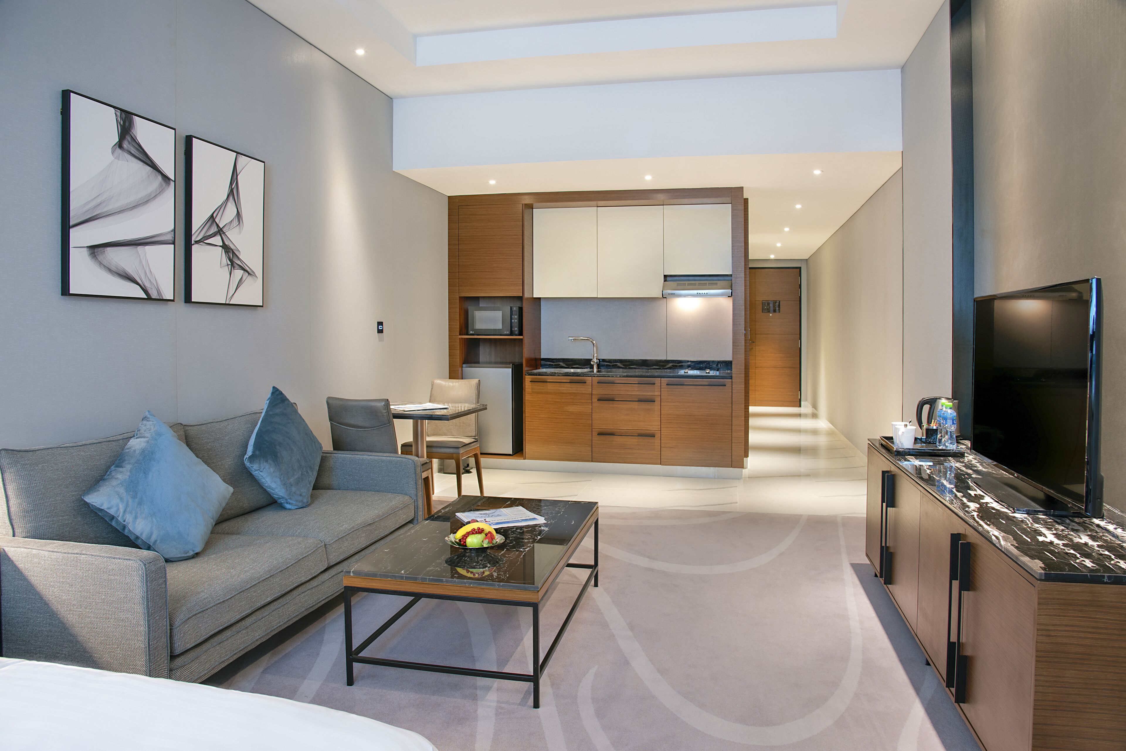 Un salon moderne avec un canapé, une table basse et une œuvre d’art au mur. On aperçoit une cuisine à l’arrière-plan, avec des meubles en bois et un téléviseur.
