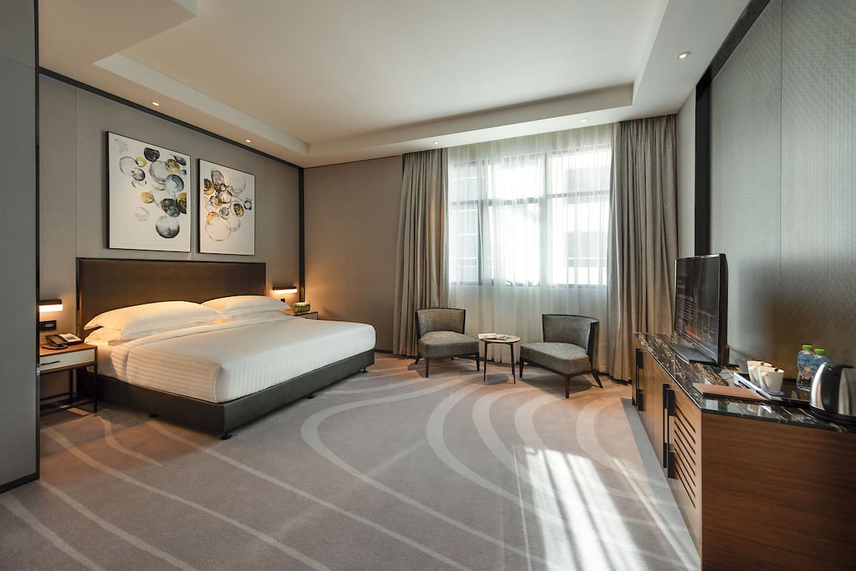 Une chambre d’hôtel moderne avec un grand lit, deux fauteuils, une petite table, un téléviseur et des œuvres d’art accrochées au mur.