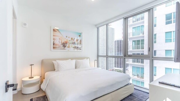 A modern hotel room with a large bed, a bedside table with a lamp, a painting on the wall, and a view of the city through large windows. - Downtown Stay | Restaurant, Gym, Pool & CoWork! (Miami)