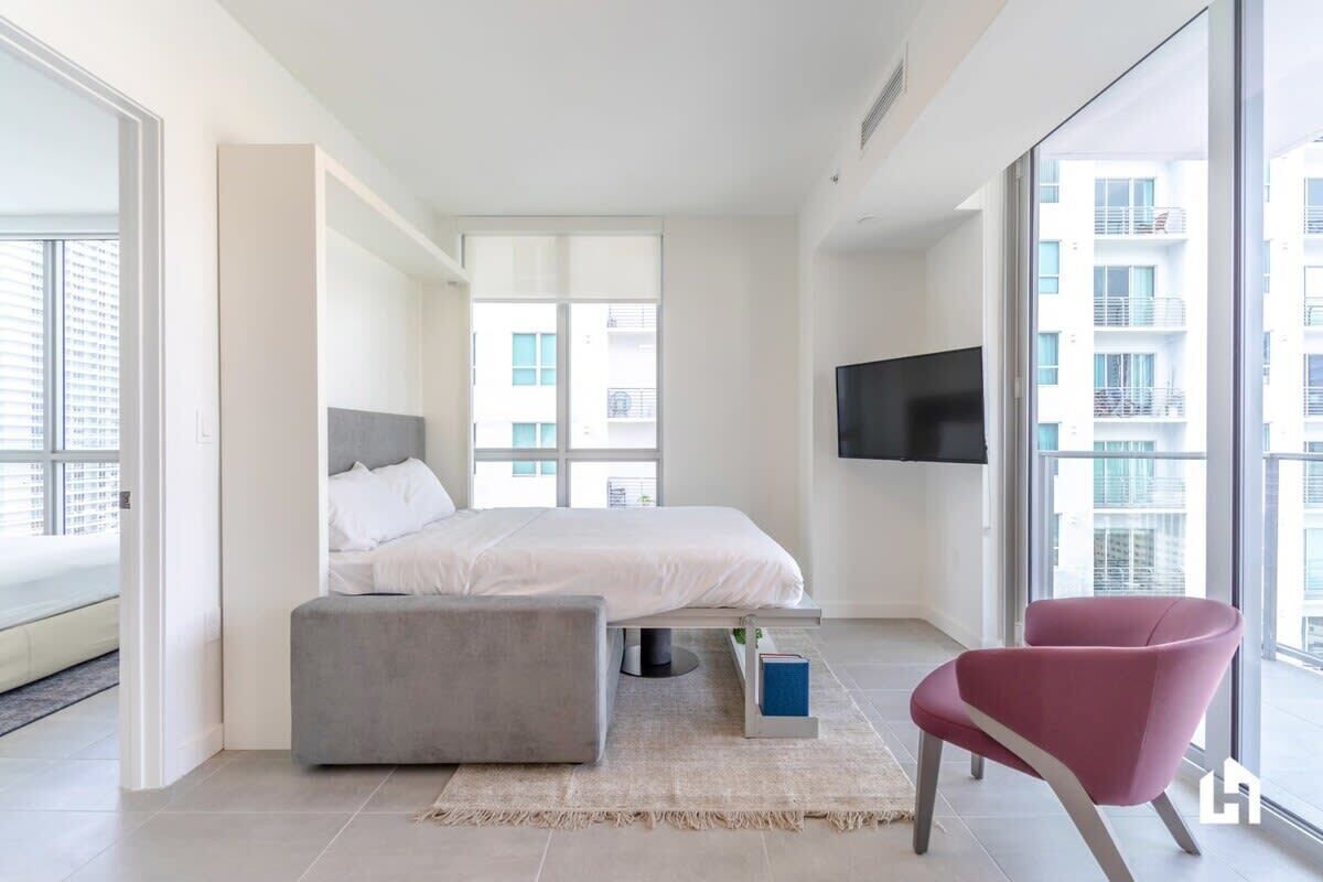 A modern hotel room with a bed, a grey ottoman, a red chair, and a TV on the wall.