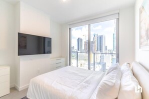 A modern bedroom with a large window, a flat-screen TV mounted on the wall, and a bed with white bedding.