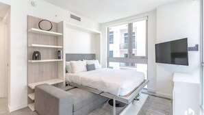 A modern bedroom with a bed, built-in shelving, a TV, and a window. - Downtown Condo with City Views, Pool, Near Bayside (Miami)