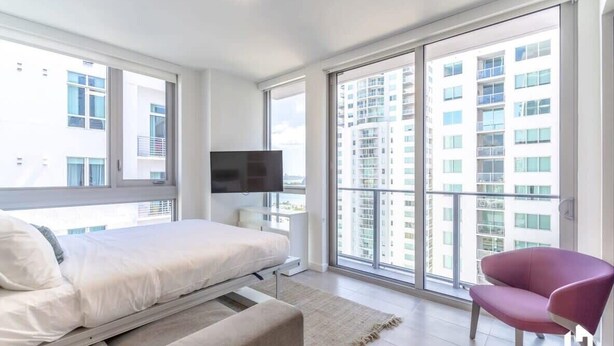 A modern bedroom with a bed, a TV, and a large window with a city view. - Downtown Condo with City Views, Pool, Near Bayside (Miami)