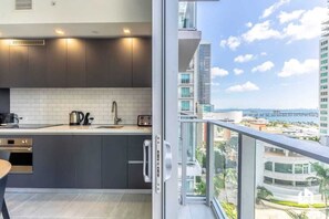 A modern kitchen with dark cabinets, white tiles, and a view of a cityscape through a balcony.