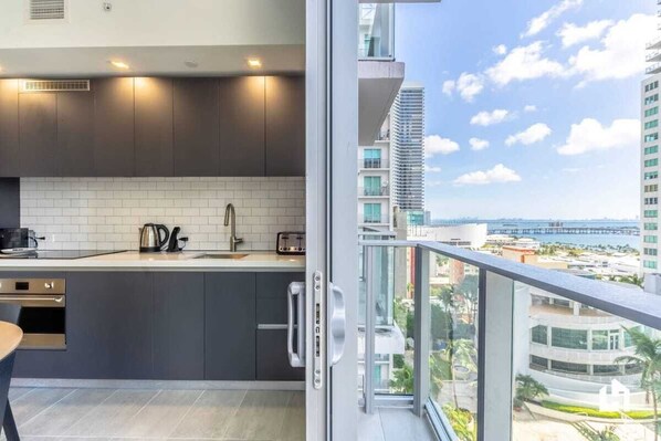 A modern kitchen with dark cabinets, white tiles, and a view of a cityscape through a balcony.