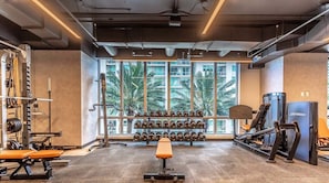 Un gimnasio con equipo de levantamiento de pesas, una banca y vistas a palmeras a través de grandes ventanales.