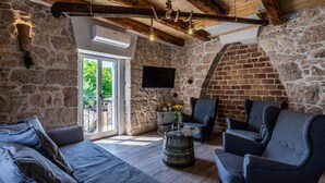 Une pièce rustique aux murs de pierre, avec un plafond en bois, un canapé, deux fauteuils, une petite table surmontée d’un tonneau et un téléviseur fixé au mur.