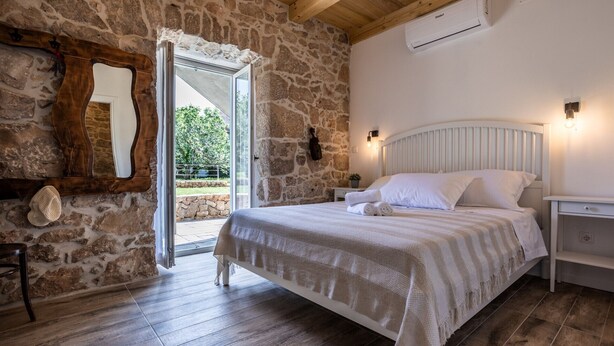 Une chambre avec un mur en pierre, un plafond en bois, un lit avec du linge de lit blanc, une table de chevet, un miroir et une vue sur l’extérieur.