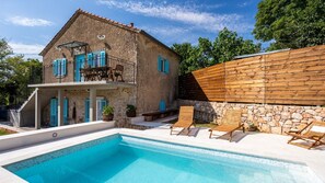 Una casa de dos pisos con piscina, muros de piedra y terraza de madera.