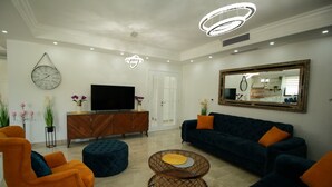 A modern living room with a dark blue sofa, orange armchairs, a wooden cabinet, a flat-screen TV, and a round coffee table.