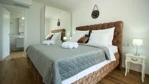 A neatly made bed with a grey quilt, white pillows, and rolled towels. There is a bedside table with a lamp and a small drawer. The room has wooden flooring and a large mirror on the wall.